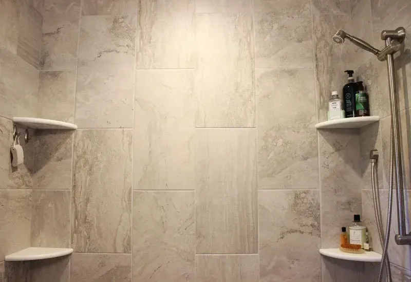 Beige tiled shower wall with built-in corner shelves holding bath products and shower fixtures on both sides  In Bucks County PA