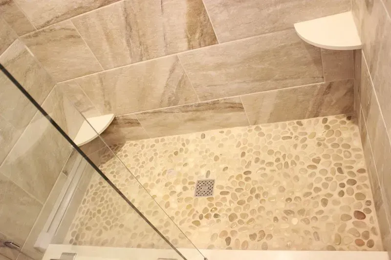 Walk-in shower with beige stone tile walls, pebble floor, and glass shower enclosure  In Bucks County PA