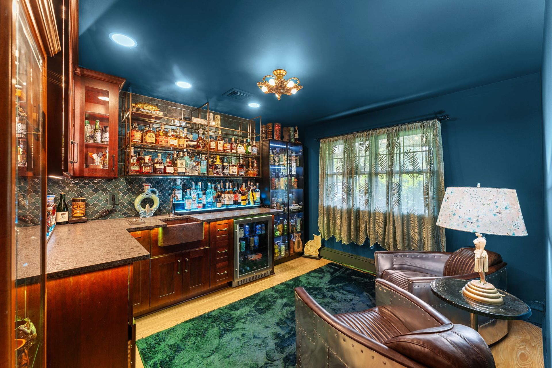 Cozy blue home bar with wood cabinets, glass shelves, and a leather chair by a window In Bucks County PA