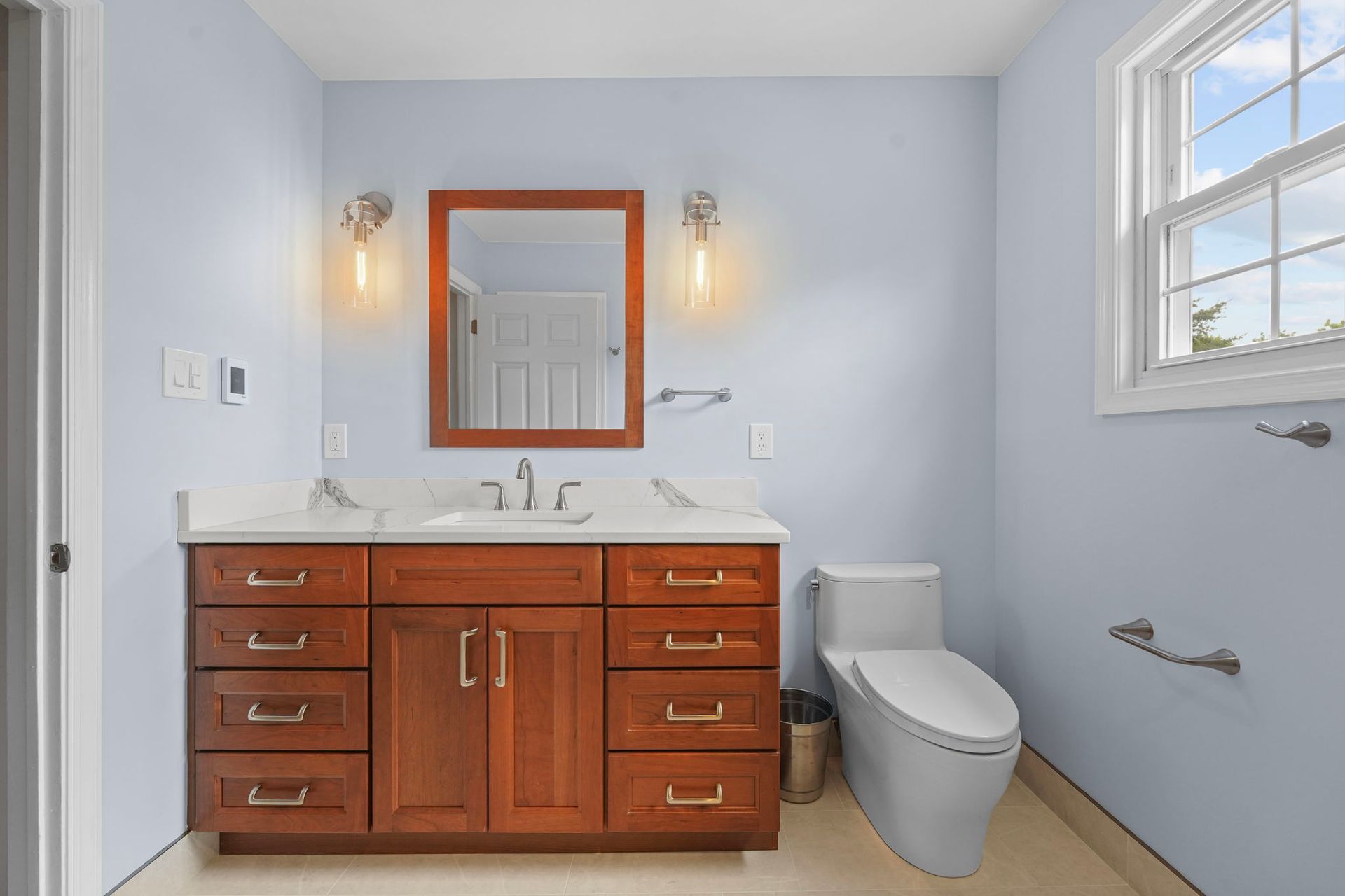 Modern bathroom with wood vanity, mirror, wall sconces, and toilet by a window  In Bucks County PA