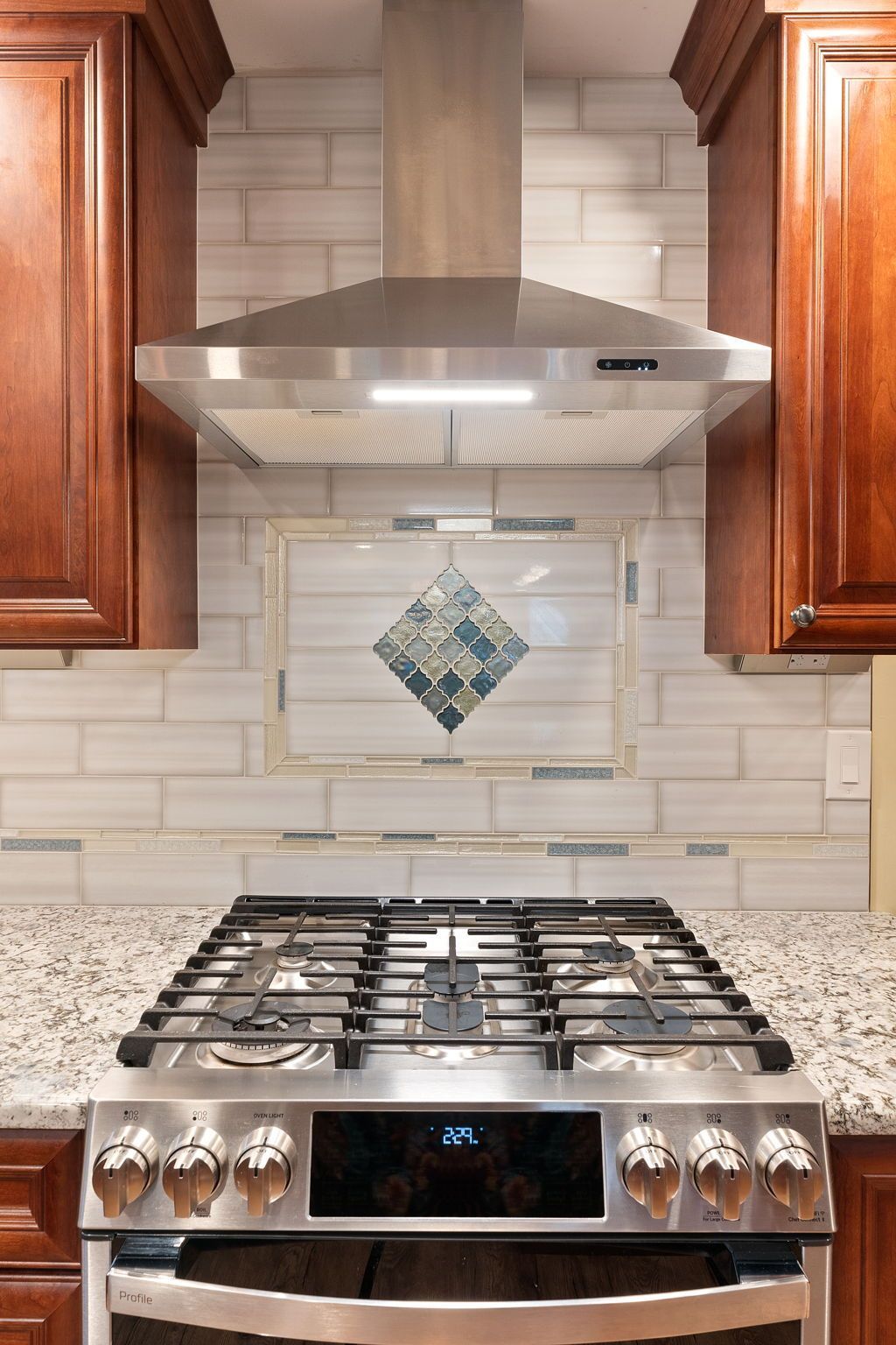 Stainless steel gas range beneath a hood in a kitchen with wood cabinets and tiled backsplash  In Bucks County PA