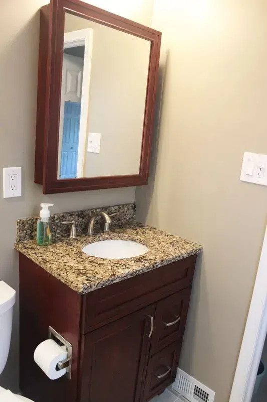 Small bathroom vanity with brown cabinet, granite countertop, oval sink, and wall mirror near a toilet paper holder  In Bucks County PA