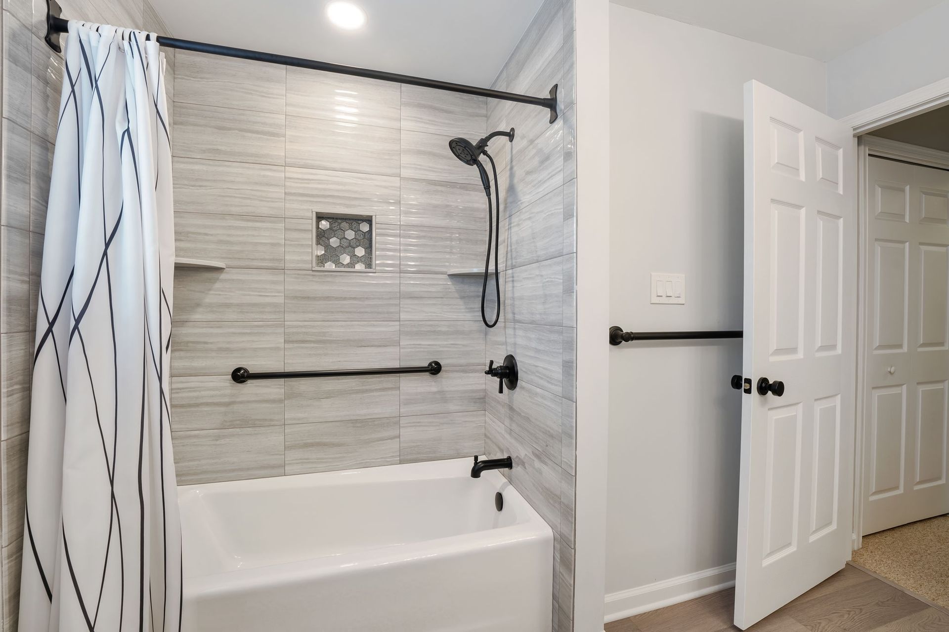 Bright bathroom with a tiled shower-tub, black grab bar, showerhead, and open door to the hall In Bucks County PA