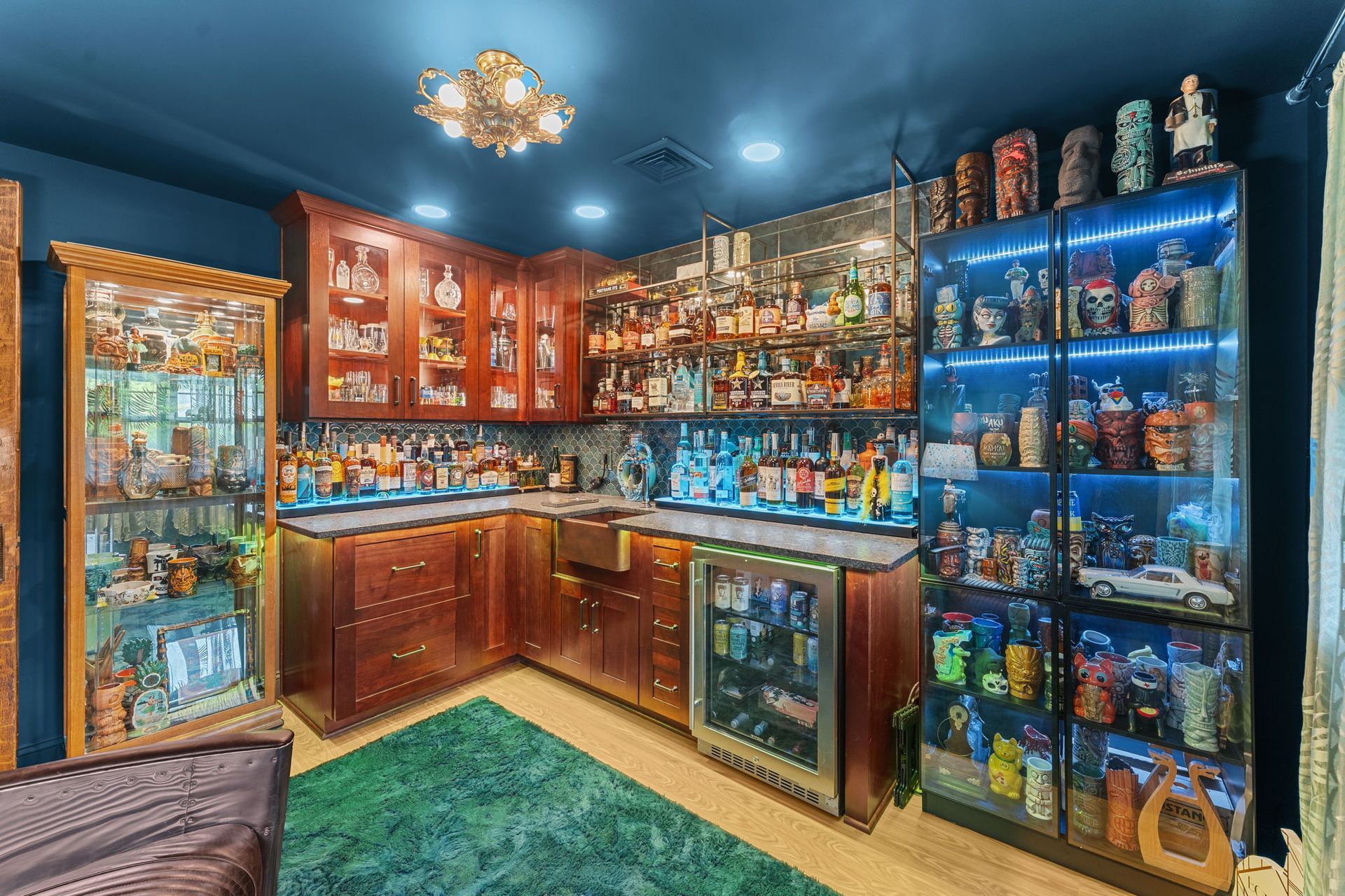 Blue home bar with wooden cabinets, glass shelves, and a green rug in a cozy basement room In Bucks County PA