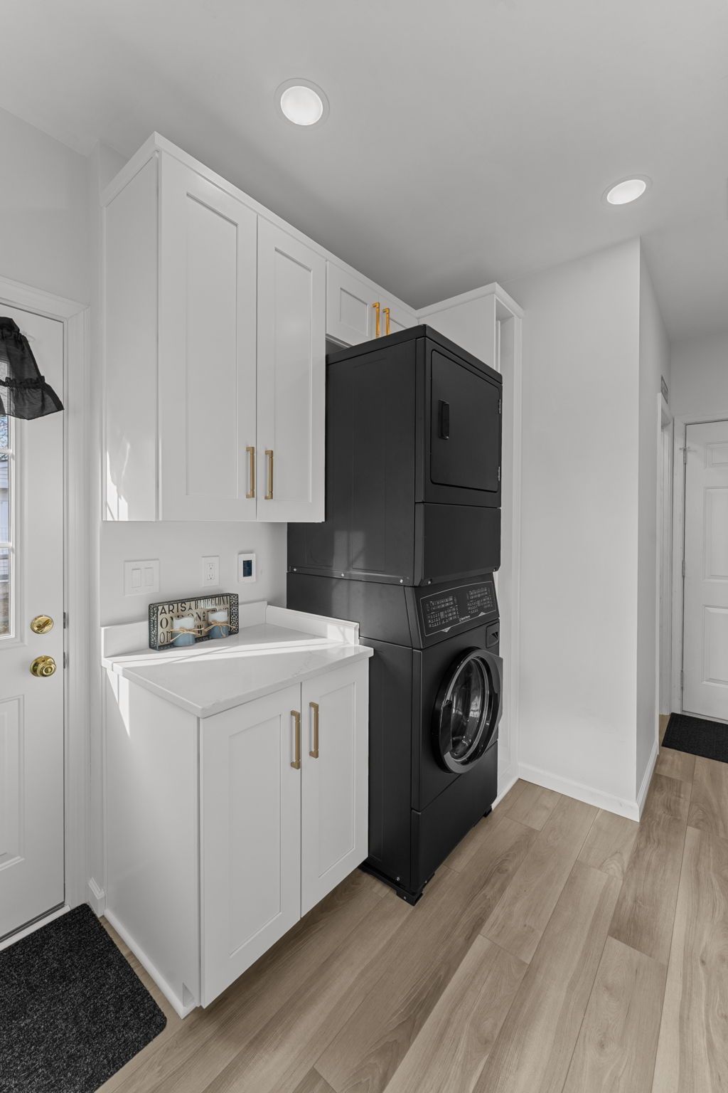 Small laundry nook with stacked black washer and dryer beside white cabinets and a sink on light wood flooring In Bucks County PA