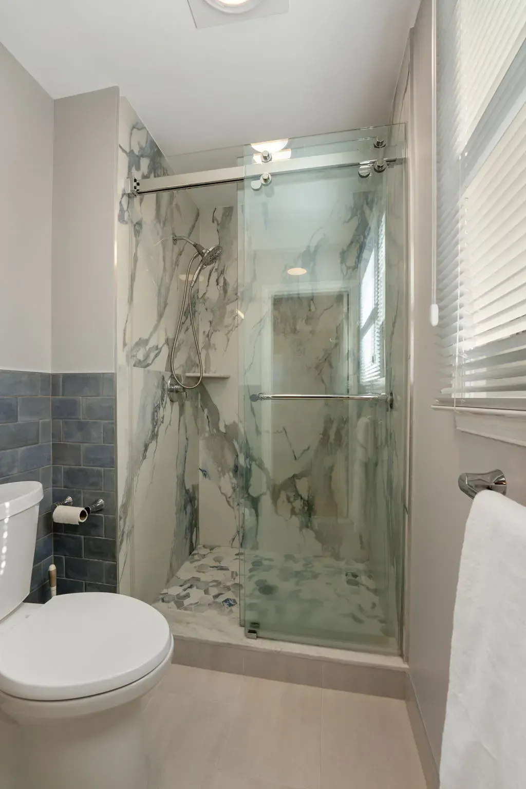 Modern bathroom with glass shower, marble-pattern tile, toilet, sink, and window blinds  In Bucks County PA