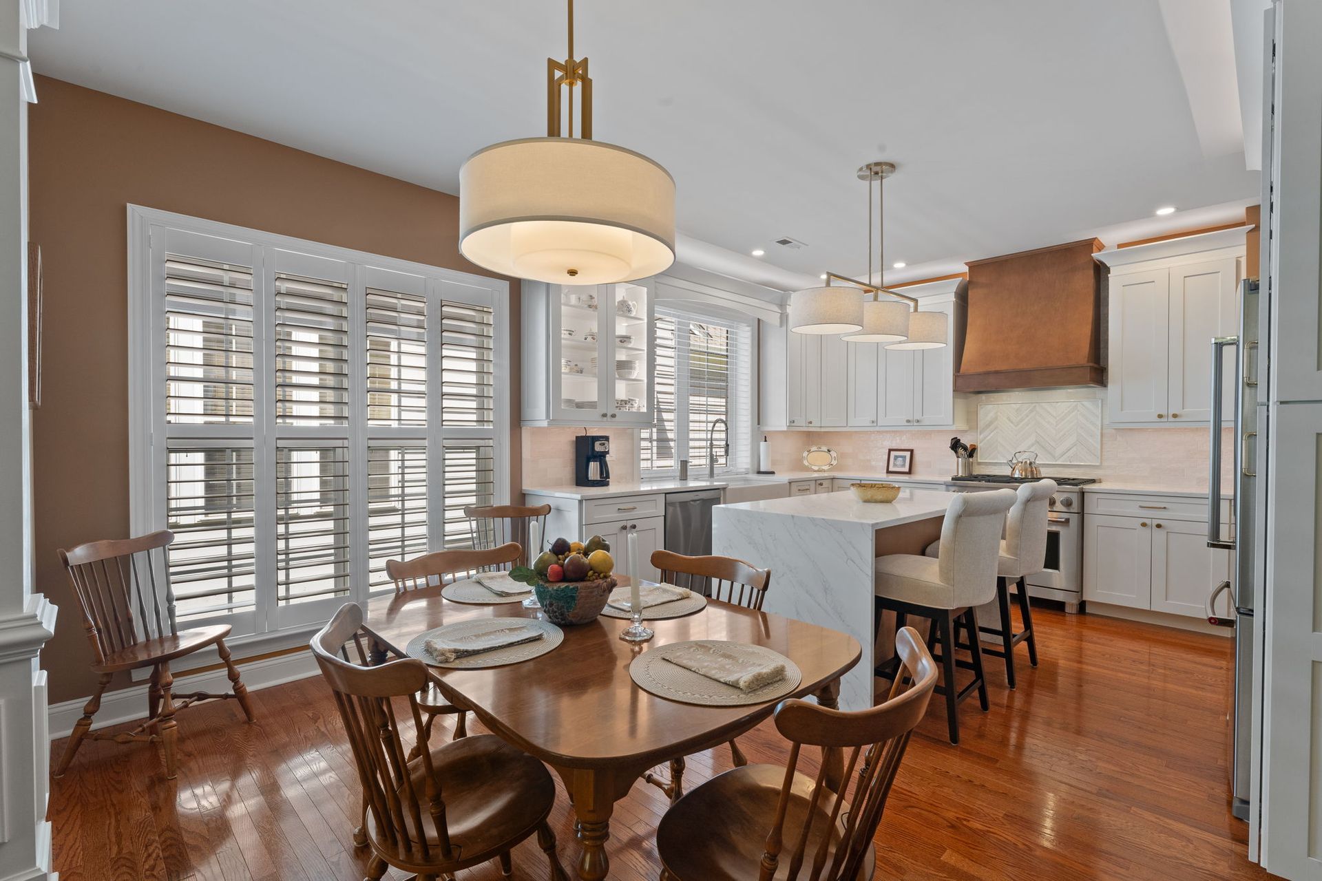 Bright kitchen with white cabinets, wood floors, island seating, and a round dining table by shuttered windows In Bucks Country PA