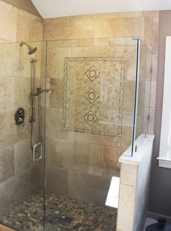 Walk-in shower with beige tile walls, pebble floor, glass partition, and built-in decorative mosaic panel  In Bucks County PA