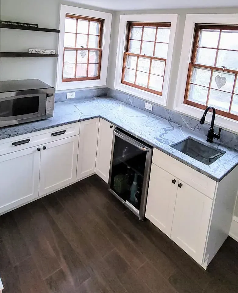 Small white kitchen with gray countertops, wood floors, three windows, microwave, sink, and cabinets In Bucks County PA