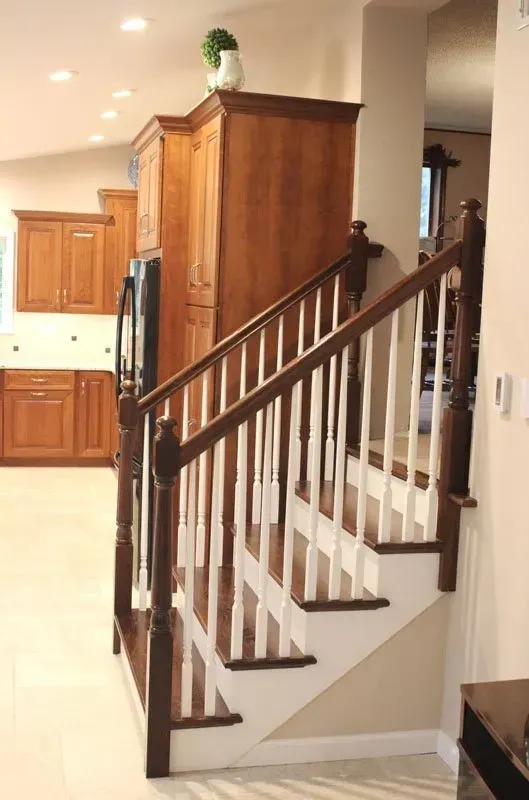 Staircase with dark wood railing beside a kitchen and hallway, with white walls and beige tile floor In Bucks County PA