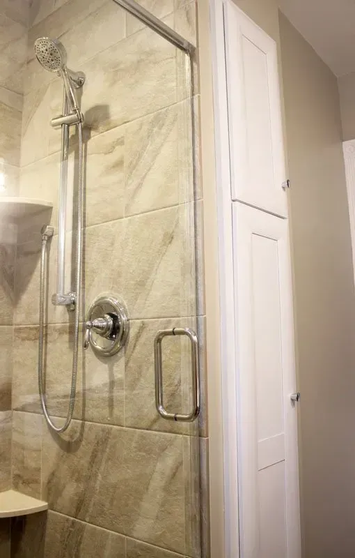 Bathroom shower with beige tile walls, glass door, and chrome grab bar near a white cabinet In Bucks Country PA