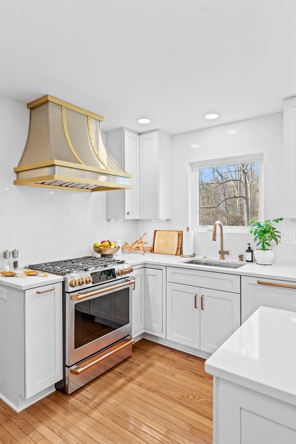 Bright white kitchen with gold range hood, stainless stove, wood floor, and window over sink In Bucks Country PA