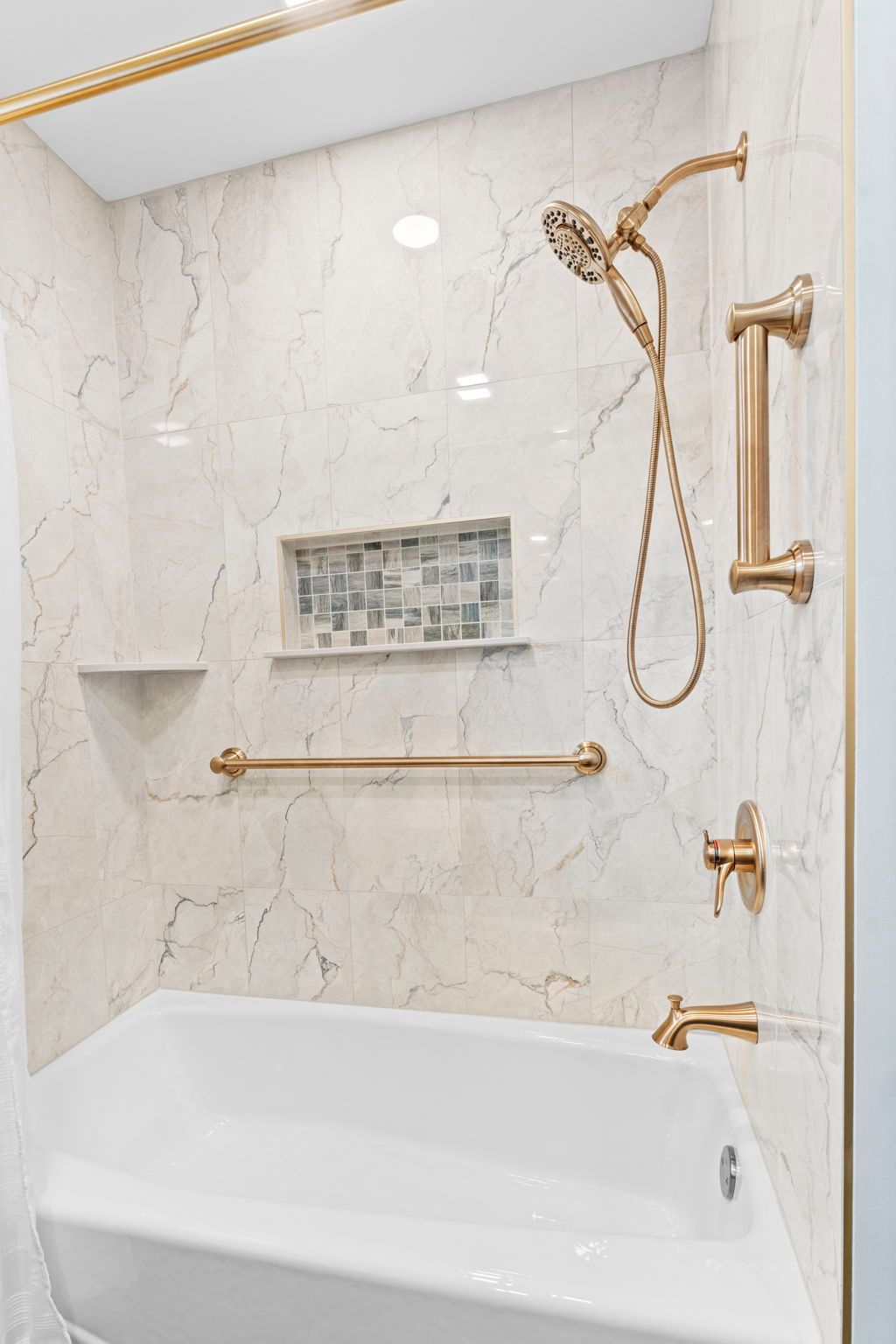 Marble tiled walk-in shower with gold fixtures, built-in shelf, grab bar, and bathtub edge in foreground In Bucks County PA