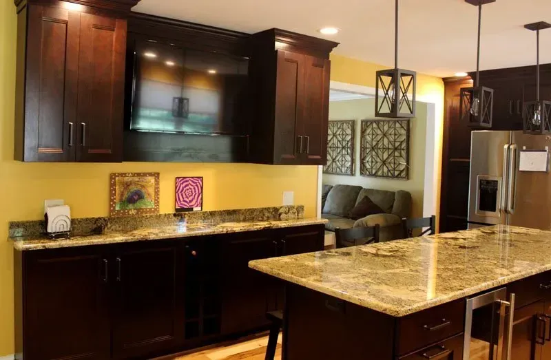 Modern kitchen with dark wood cabinets, granite island, stainless-steel fridge, and a TV above the counter In Bucks County PA