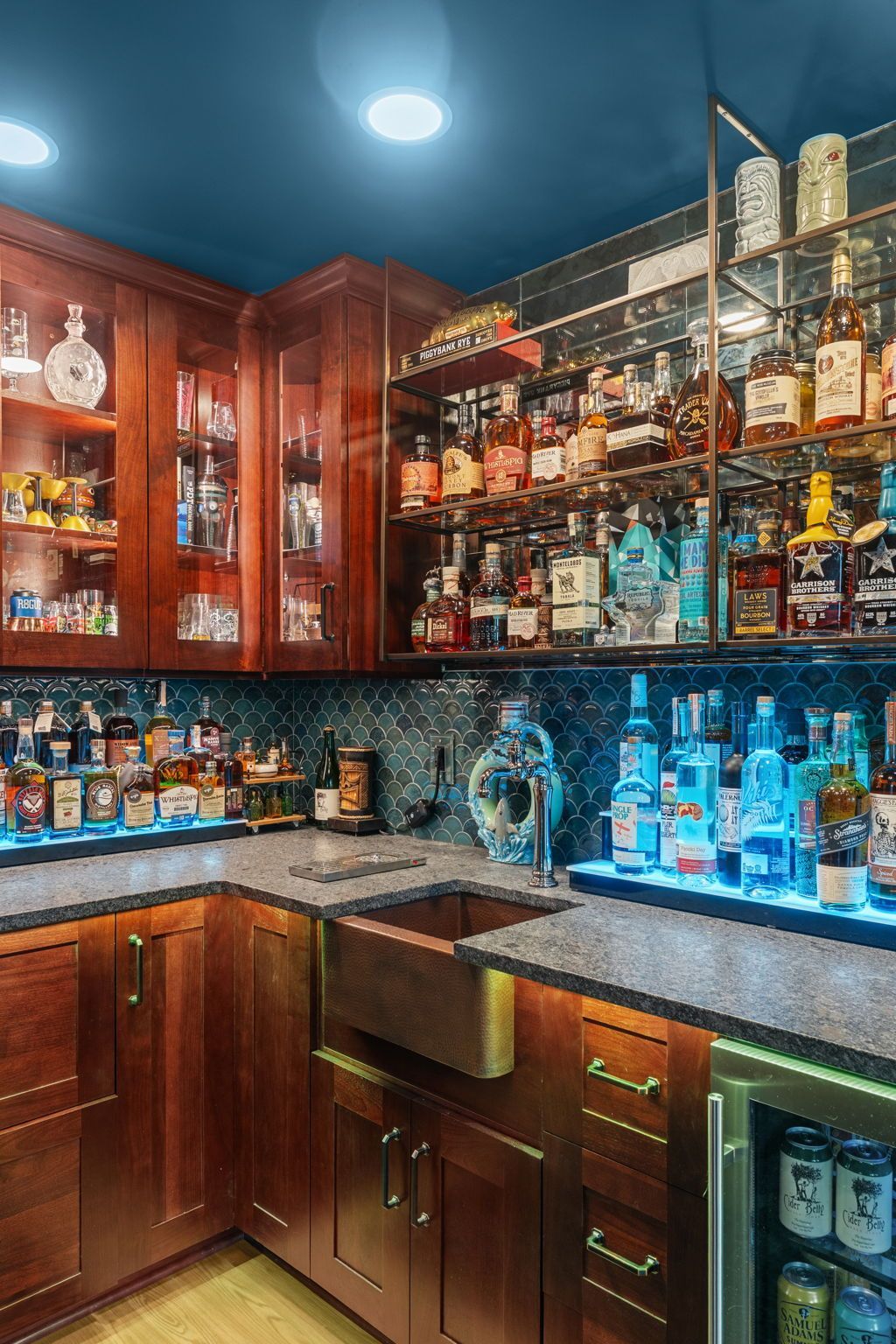 Warmly lit home bar with wood cabinets, glass shelves, and rows of liquor bottles under blue ceiling lights In Bucks County PA