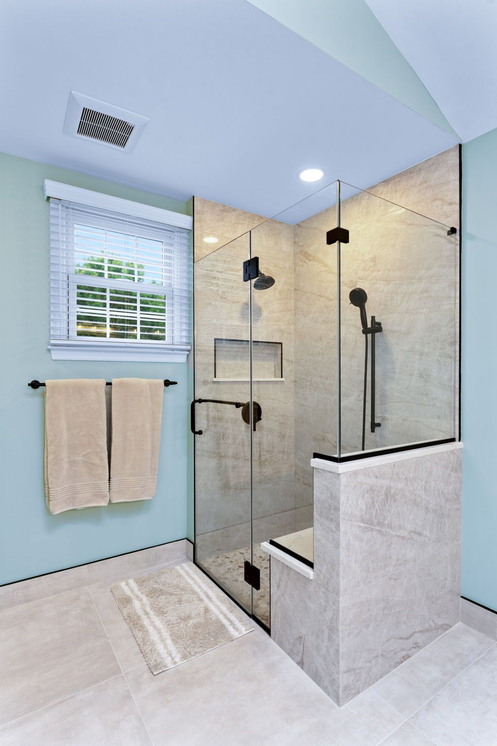 Modern walk-in shower with glass door, beige tile, black fixtures, white tub ledge, and light blue walls In Bucks County PA