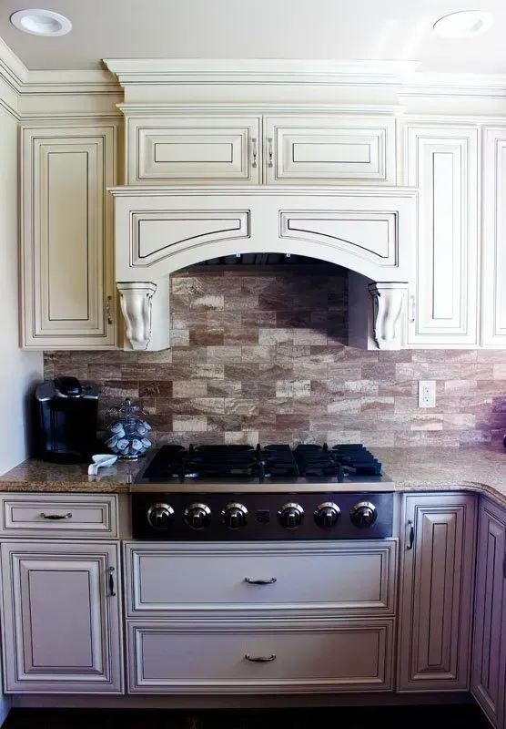 Cream kitchen with decorative cabinets, stone backsplash, and stainless stove beneath a curved hood  In Bucks County PA