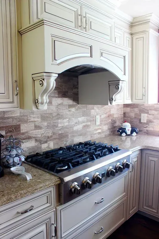 Cream-colored kitchen stove with black burners, ornate hood, and stone tile backsplash  In Bucks County PA