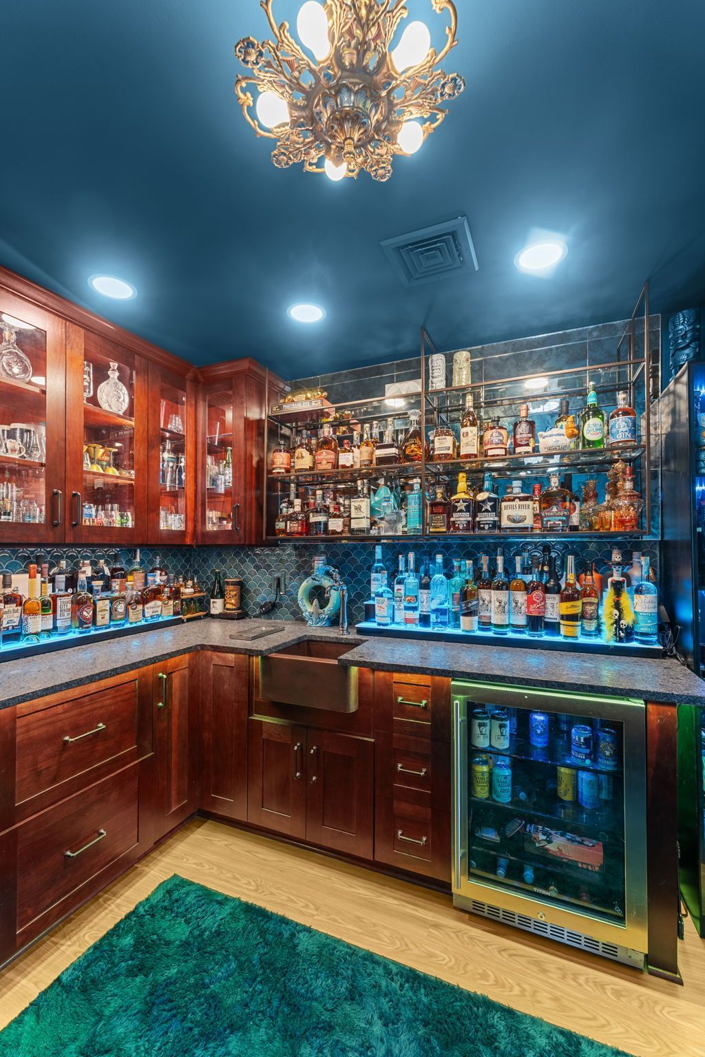 Home bar with dark wood cabinets, colorful liquor bottles, blue ceiling lights, and a small beverage fridge In Bucks County PA