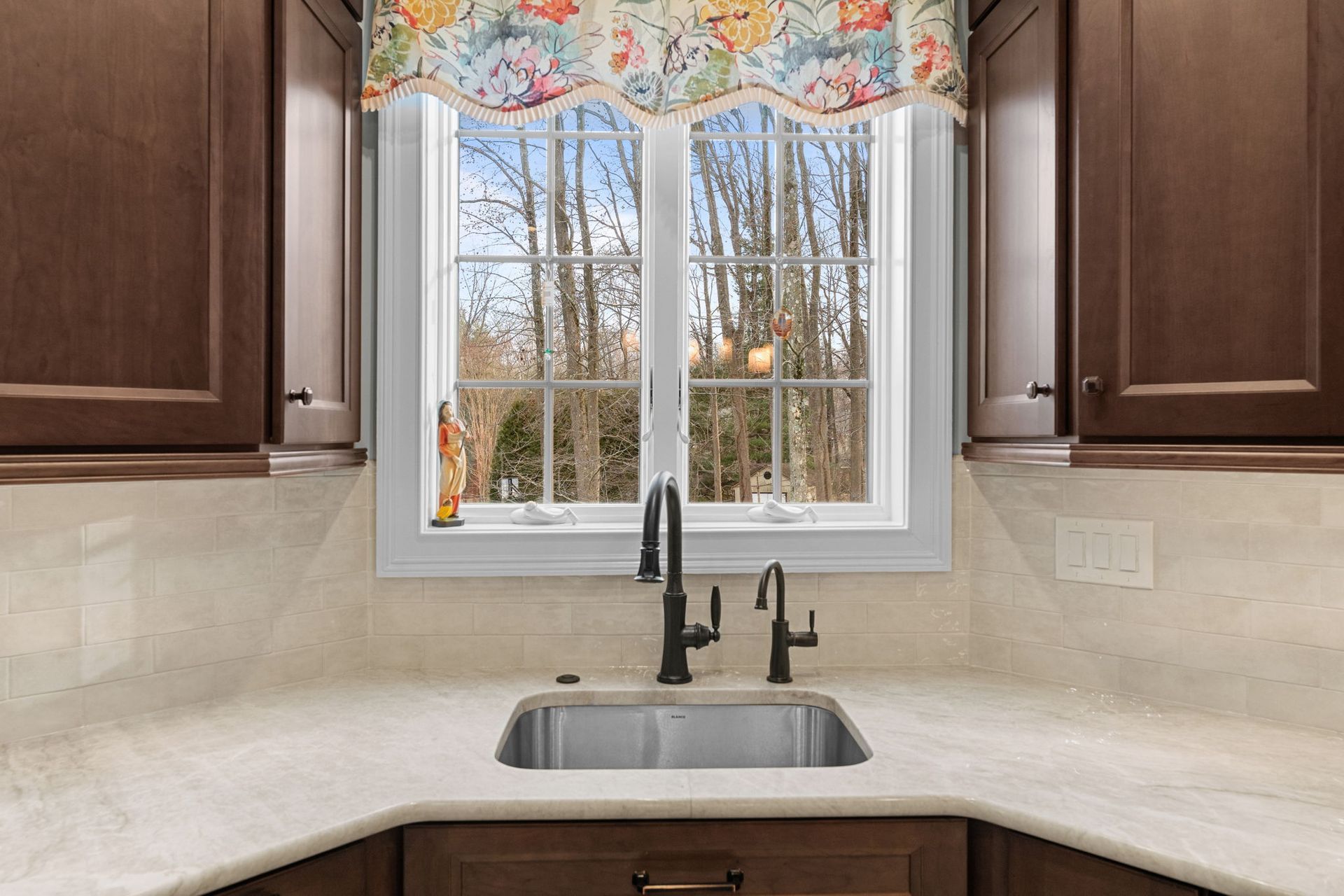 Kitchen sink beneath a window with floral curtain, dark wood cabinets, and light countertops In Bucks Country PA