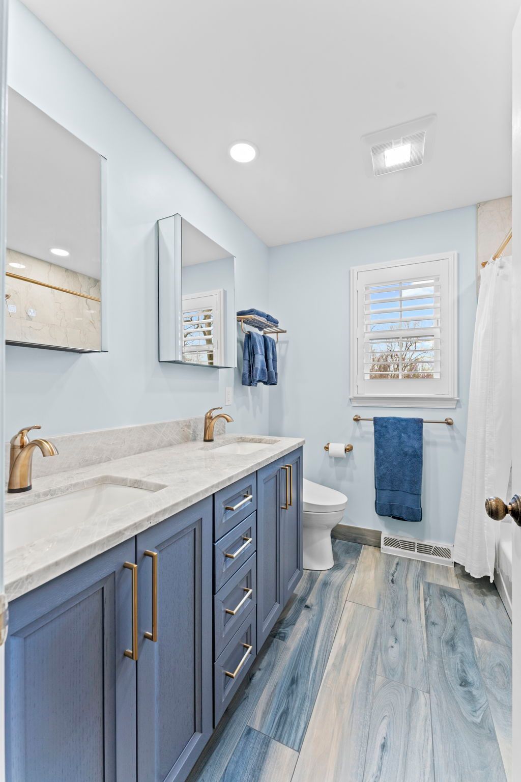 Light blue bathroom with vanity, toilet, shower curtain, and window, featuring blue-gray cabinetry and floor  In Bucks County PA