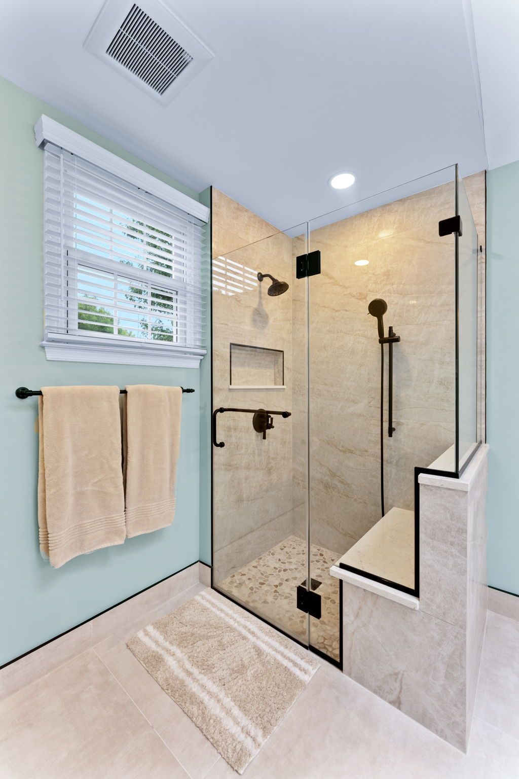 Compact tiled walk-in shower with glass door, bench, black fixtures, and a window with towel rack nearby In Bucks County PA
