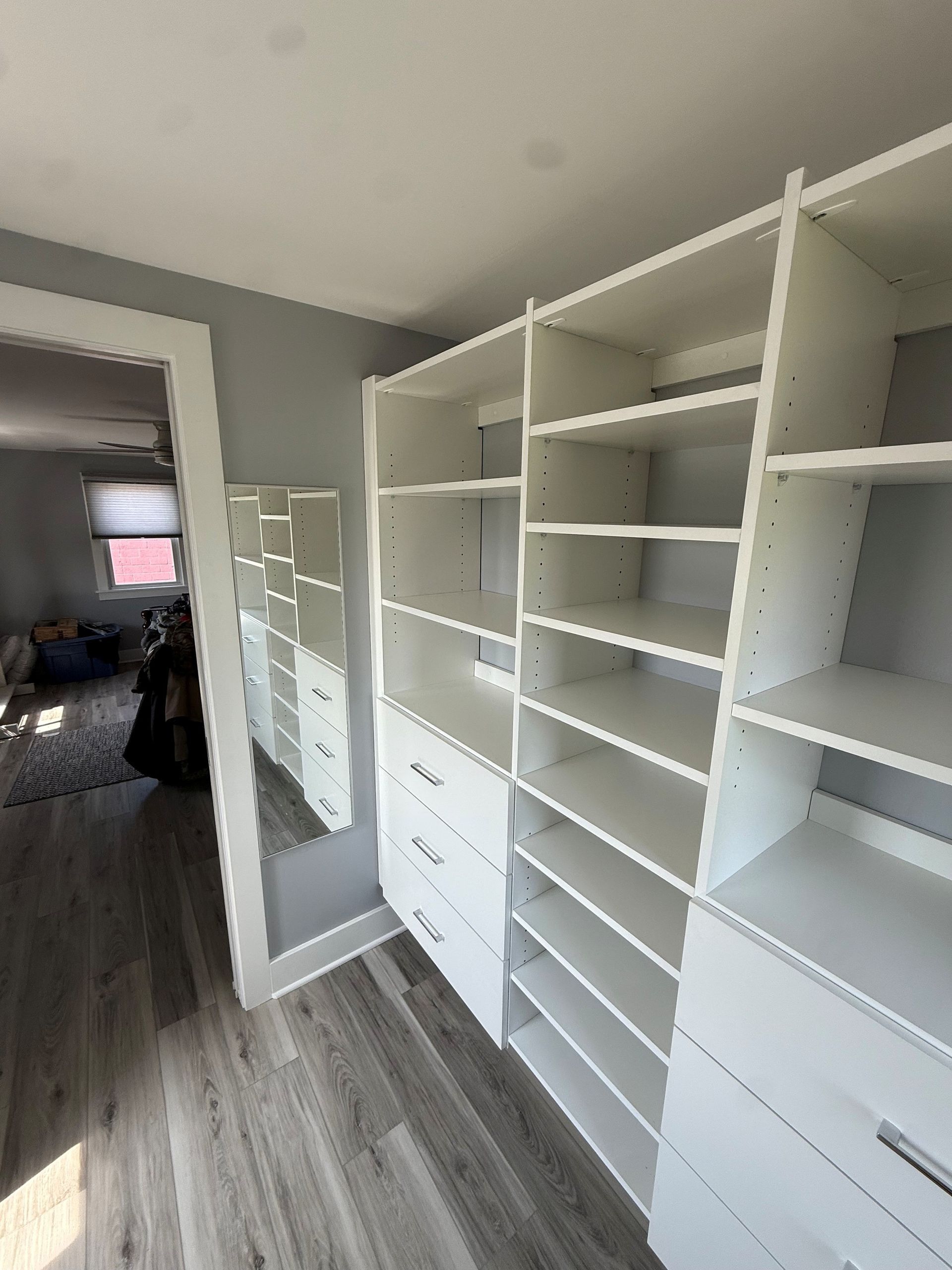 Walk-in closet with white built-in shelves, drawers, and a mirrored door on a gray wood floor In Bucks County PA