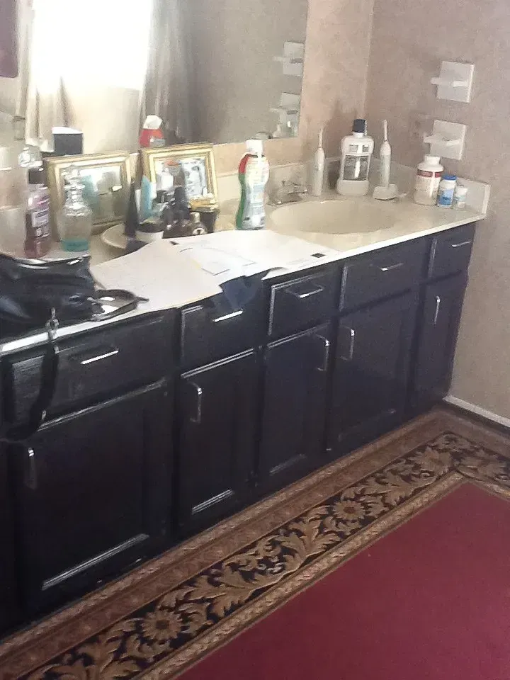 Black bathroom vanity with mirror, beige countertop, sink, toiletries, and a patterned rug on the floor  In Bucks County PA
