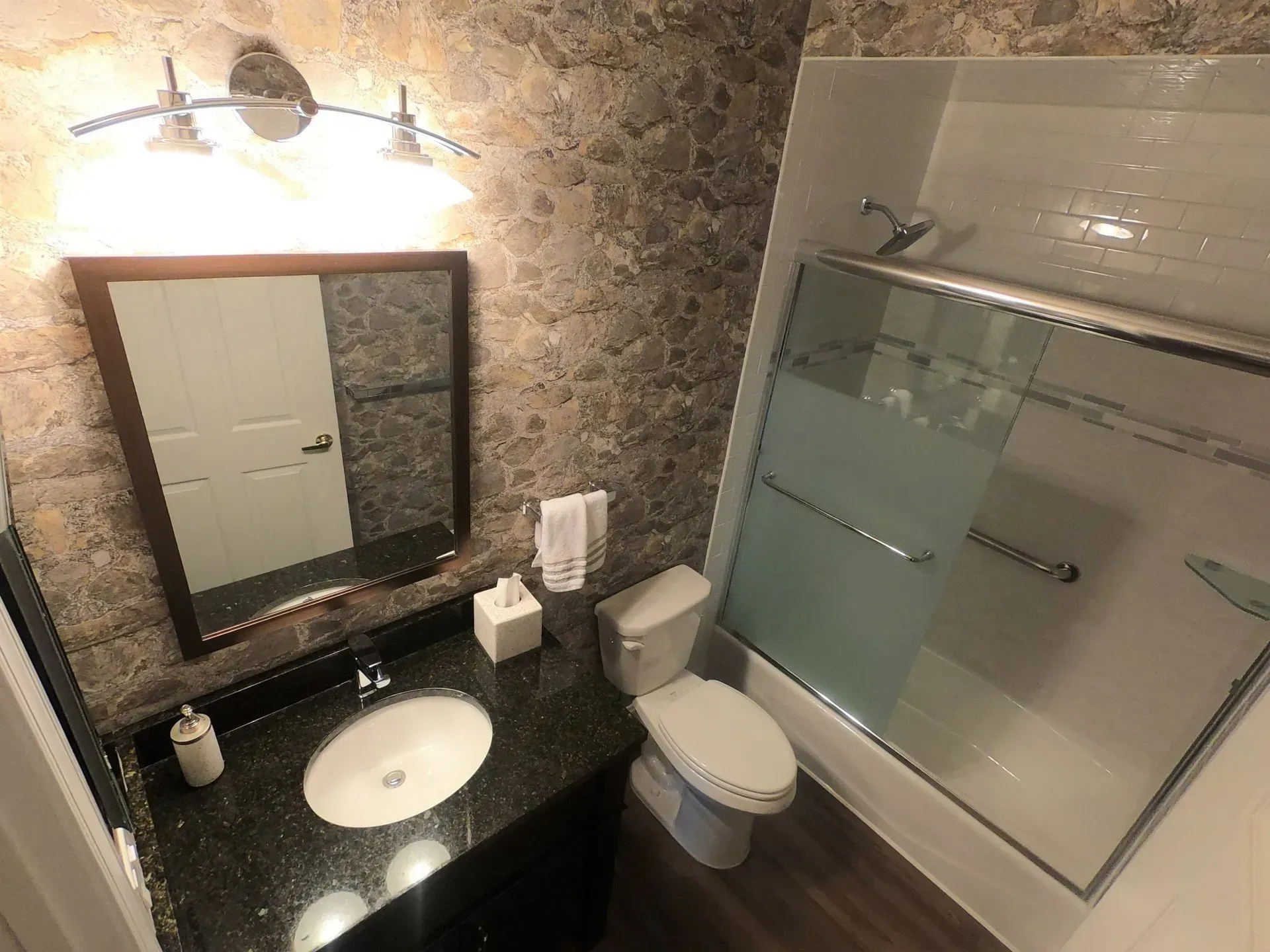 Small bathroom with stone wall, black vanity, mirror, toilet, and glass shower enclosure  In Bucks County PA
