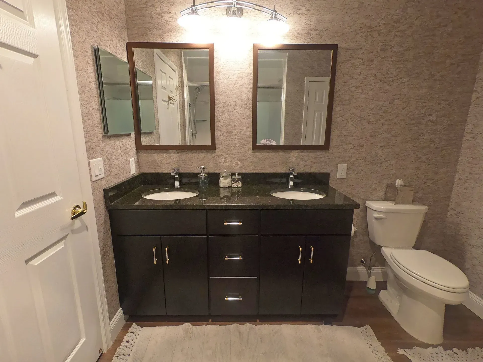 Bathroom with dark vanity, double sinks, mirrors, and a white toilet against patterned wallpaper In Bucks County PA