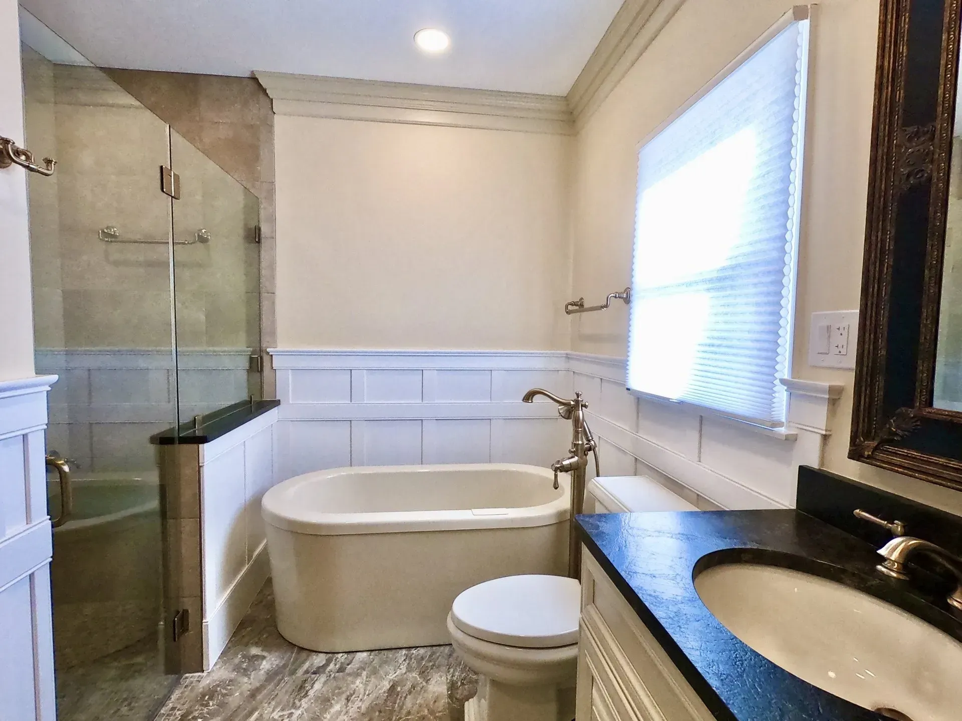 Modern bathroom with glass shower, freestanding tub, white wainscoting, and granite vanity near a window  In Bucks County PA