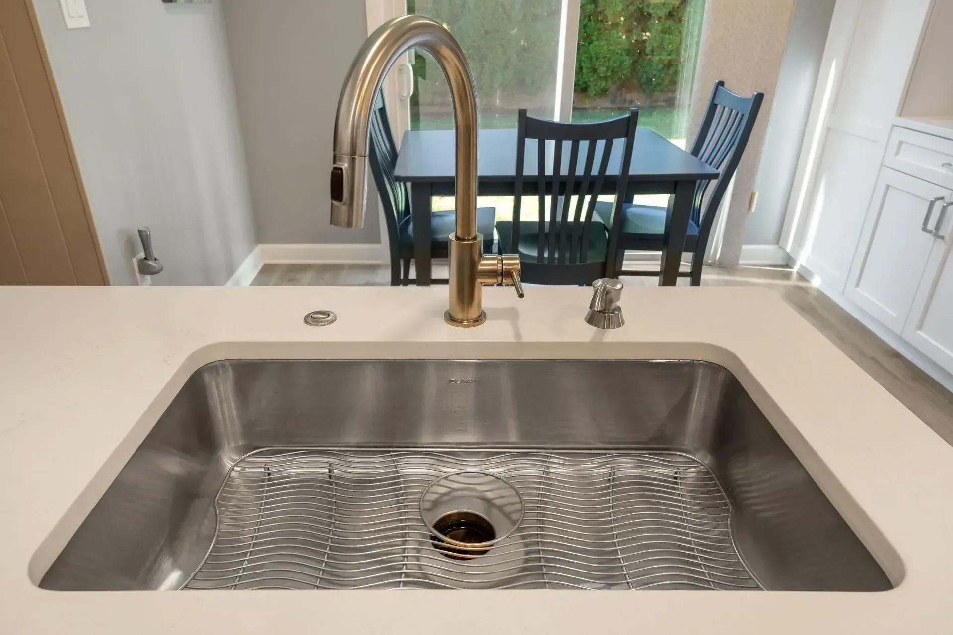 Kitchen sink with brass faucet on a white countertop, overlooking a dining table and chairs in Bucks County PA
