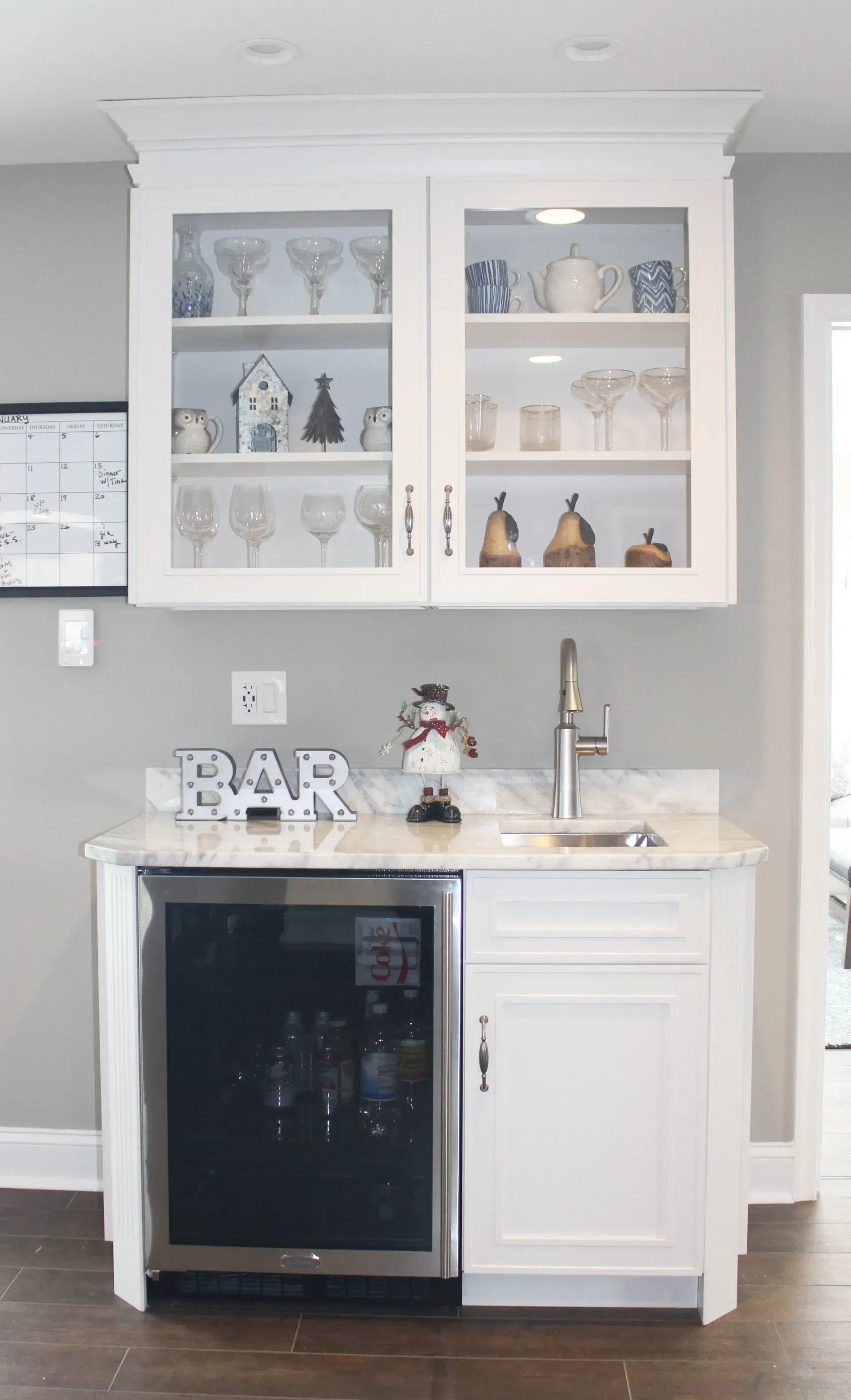 White home bar with glass cabinets, mini fridge, sink, and decorative glassware against a light gray wall  In Bucks County PA