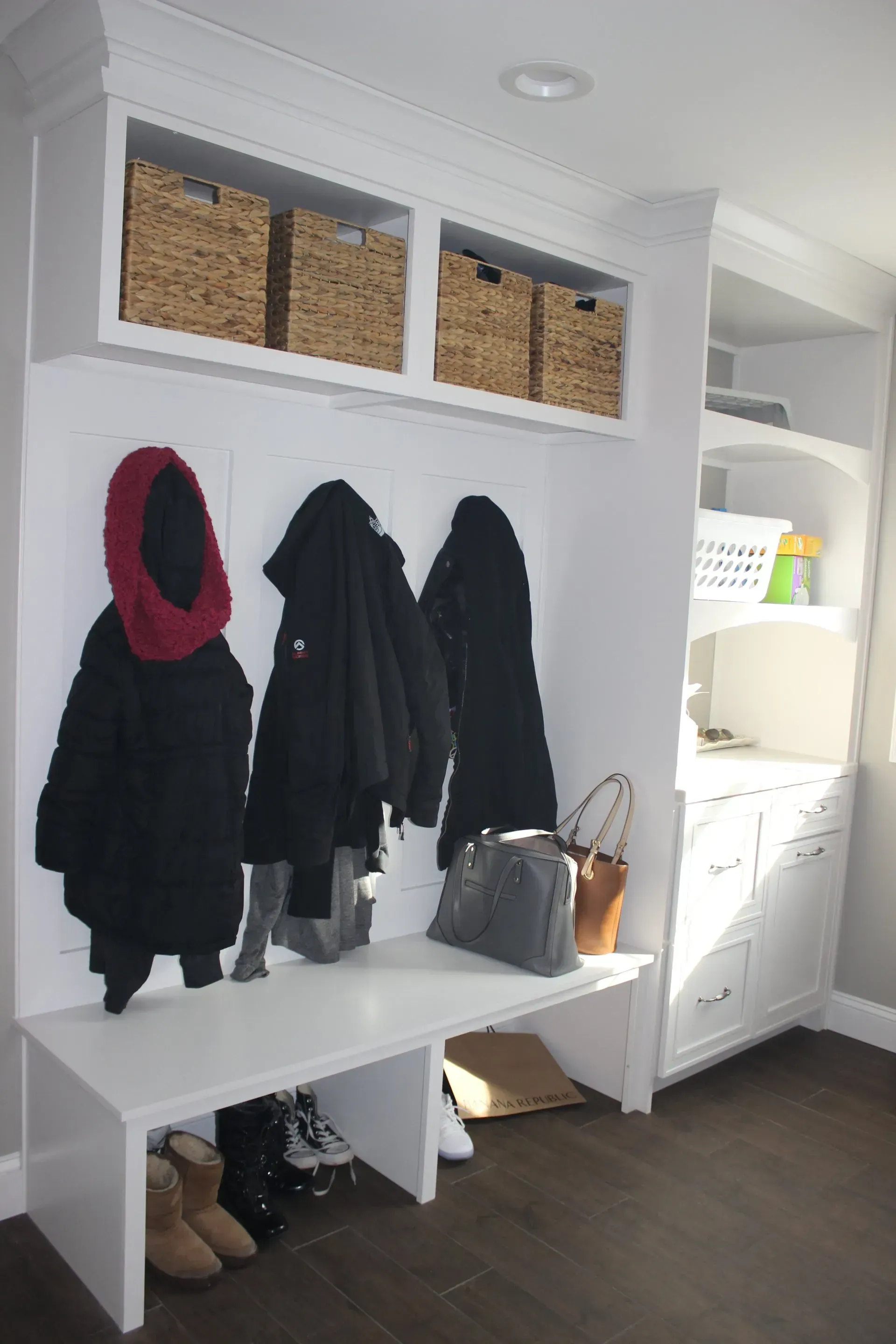 White entryway with built-in coat hooks, jackets, baskets, bench, and a nearby kitchen pass-through  In Bucks County PA