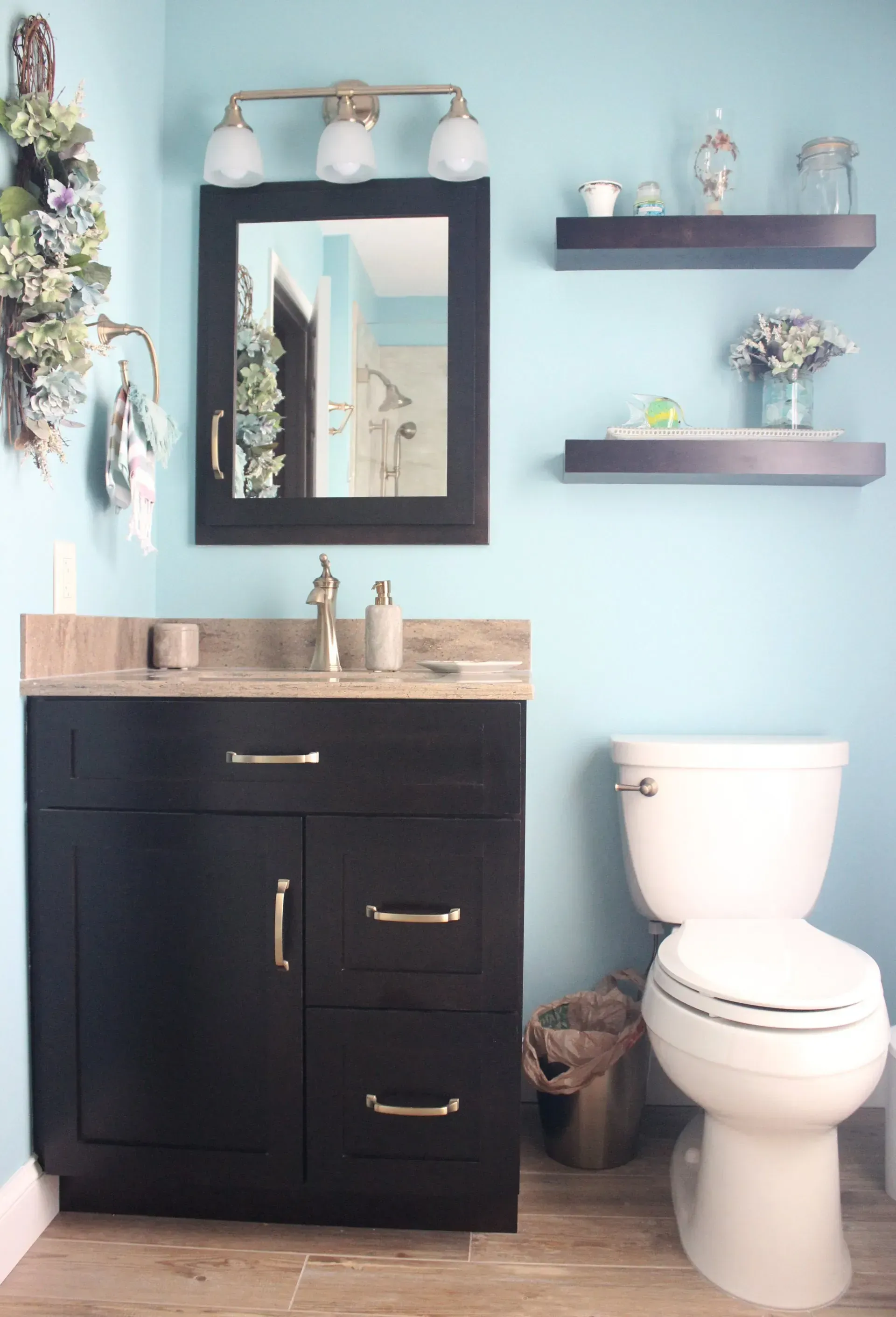 Bathroom with light blue walls, dark vanity, mirror, toilet, and two floating shelves with decor In Bucks County PA