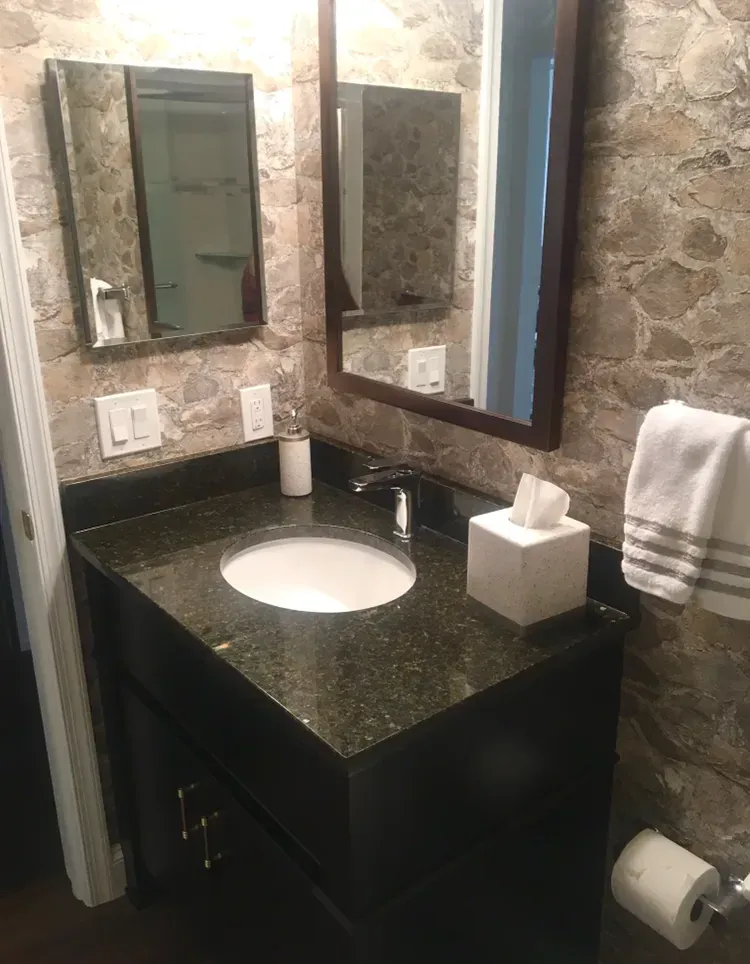 Small bathroom vanity with black countertop, round sink, wall mirror, tissue box, and towel against stone walls  In Bucks County PA