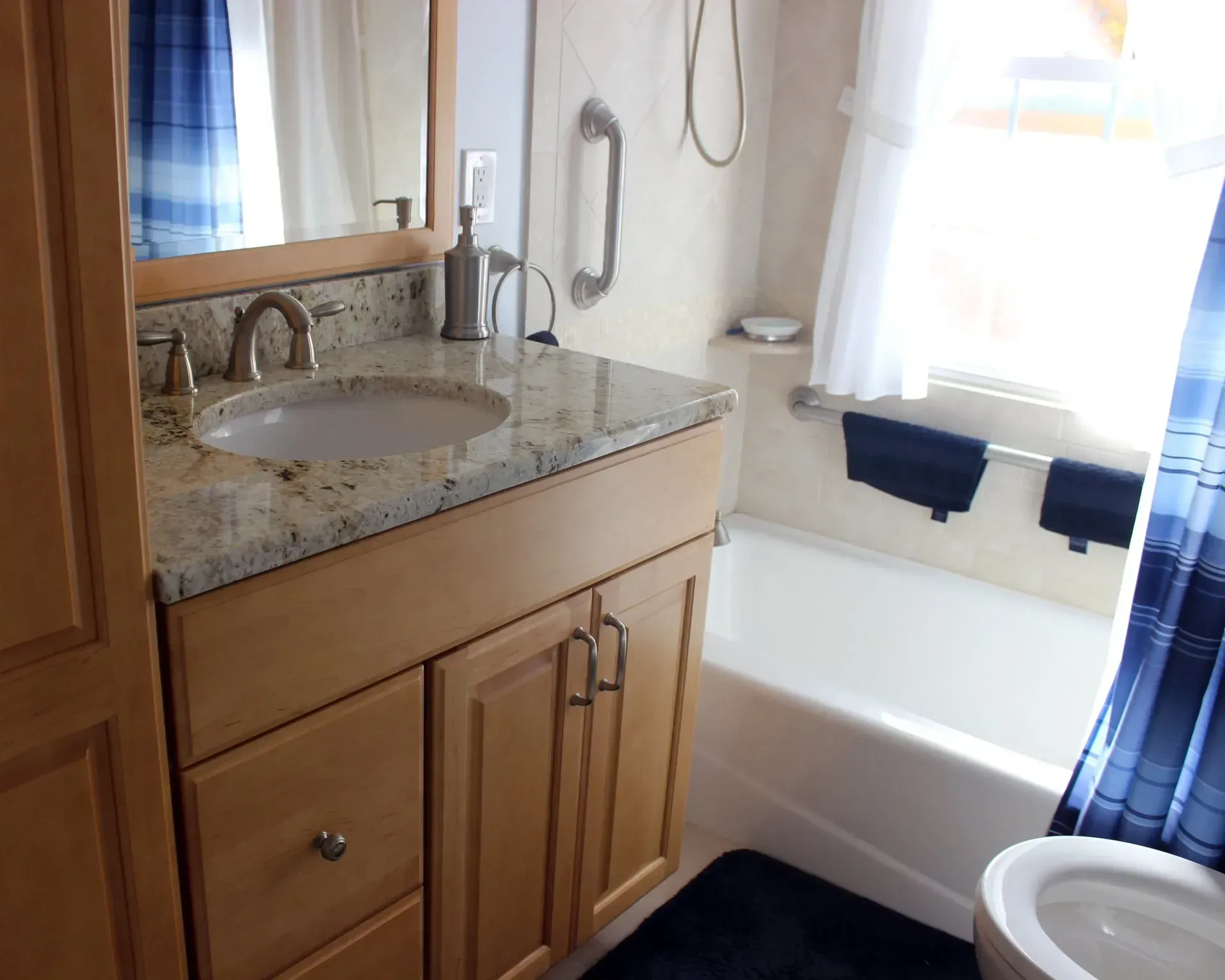 Accessible bathroom with granite vanity, sink, toilet, and tub near a window with blue curtains In Bucks County PA