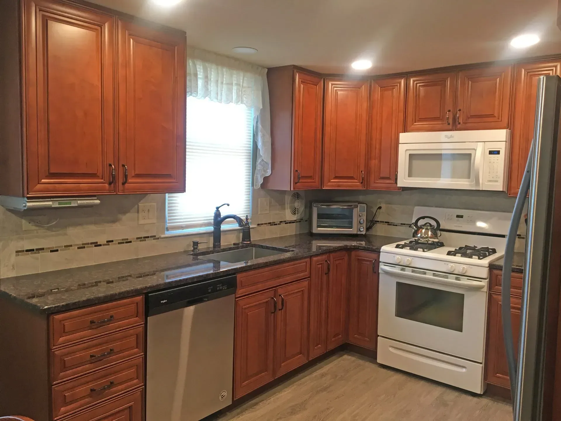 Modern kitchen with wooden cabinets, granite counters, stainless dishwasher, white stove and microwave, and a window over the sink In Bucks Country PA
