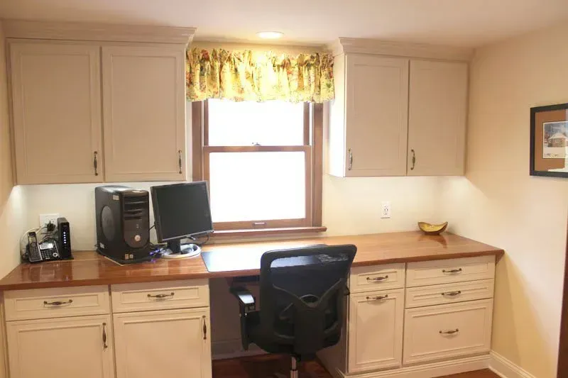 Home office nook with beige cabinets, wooden desk, computer, and black chair beneath a window with yellow valance in Bucks County PA