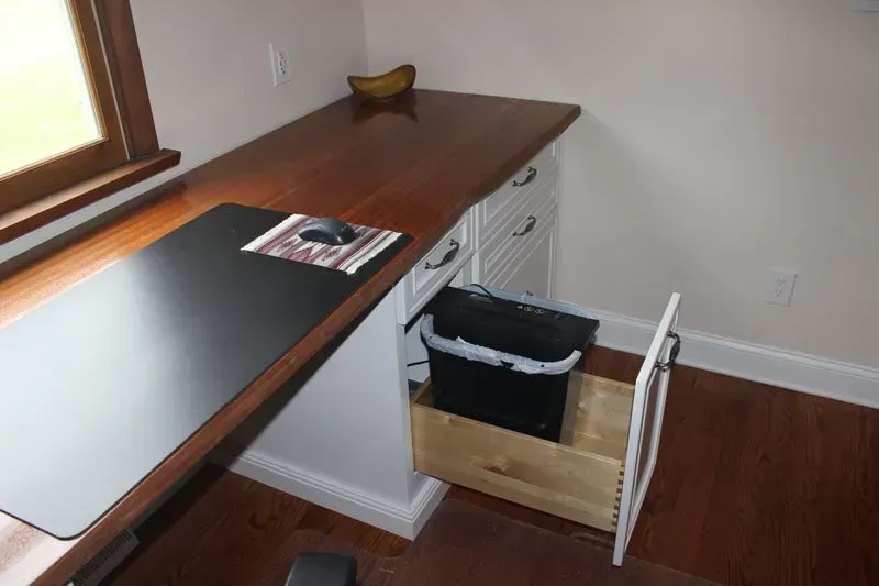 Wooden standing desk by a window with a black mat, drawer, and open cabinet holding a trash bin in Bucks County PA