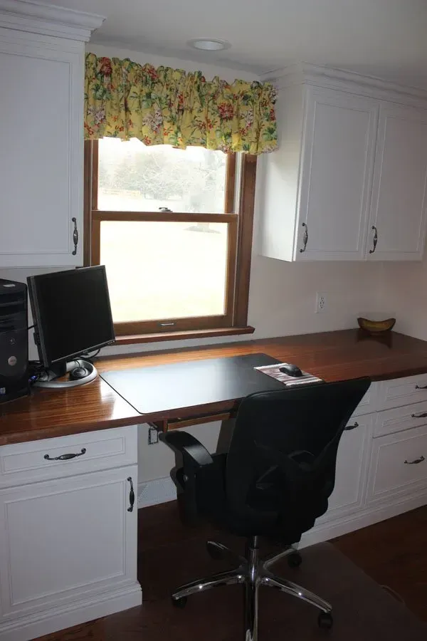Home office with white cabinets, wood desk, computer monitor, and black chair by a window with floral valance in Bucks Country PA