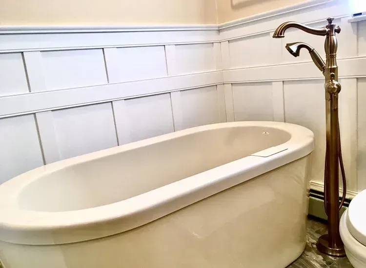 Freestanding white bathtub with brass faucet in a white paneled bathroom  In Bucks County PA