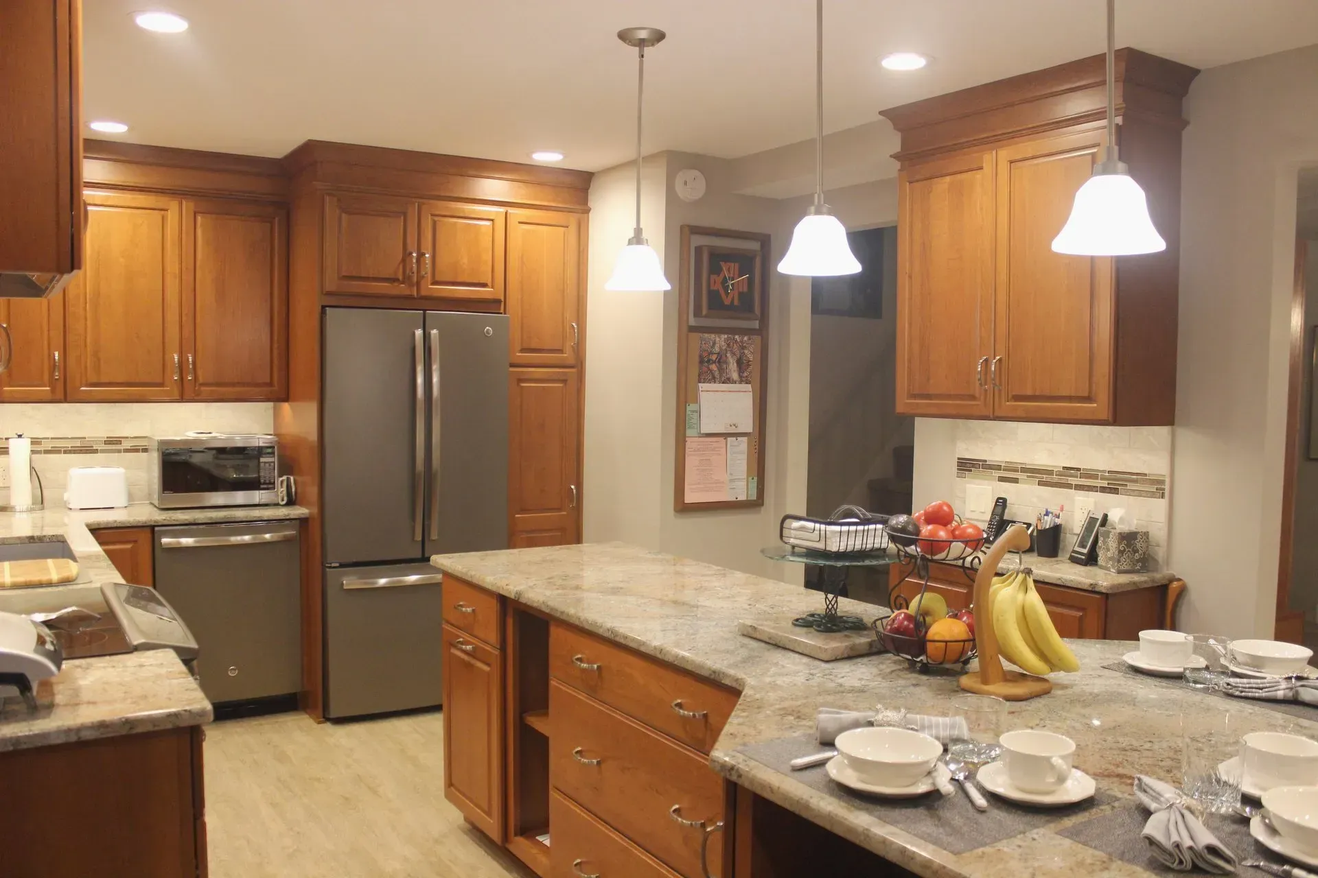 Warm kitchen with wooden cabinets, granite counters, stainless fridge, and set dining table with fruit centerpiece In Bucks County PA