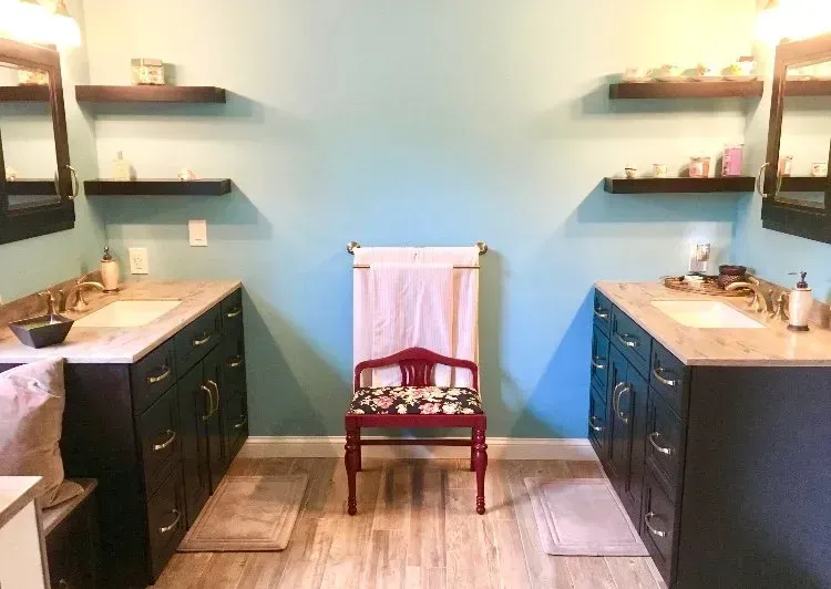 Modern double-sink bathroom with dark vanities, wall shelves, and a wooden chair centered between them  In Bucks County PA