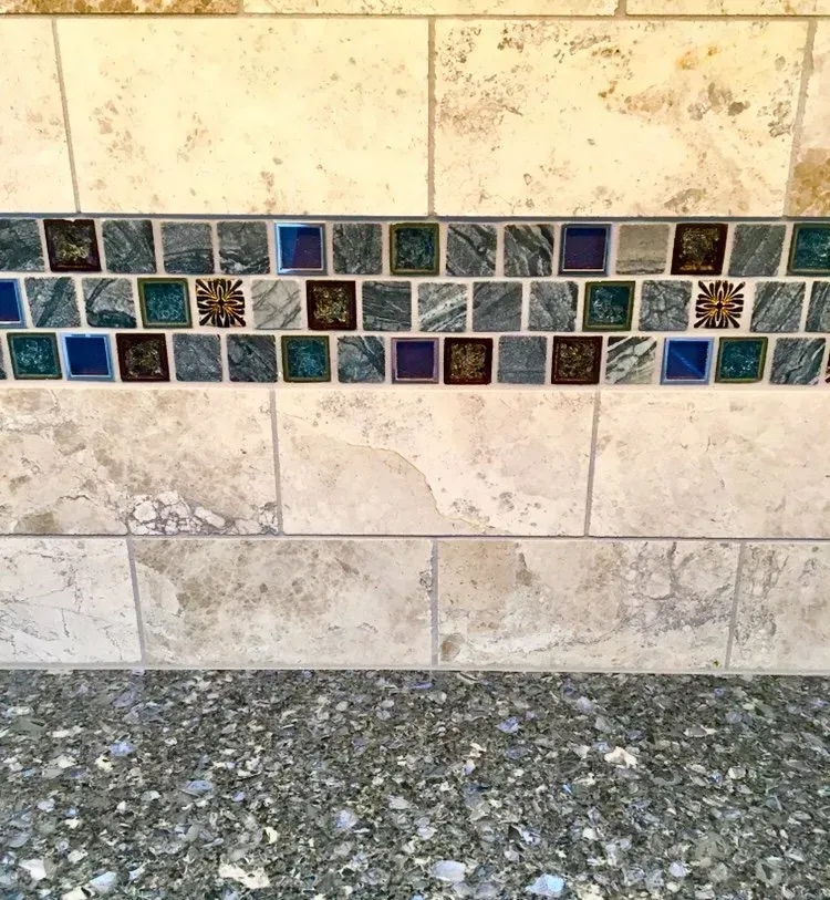 Close-up of beige stone wall with a horizontal strip of blue and green mosaic tiles above a speckled countertop In Bucks County PA