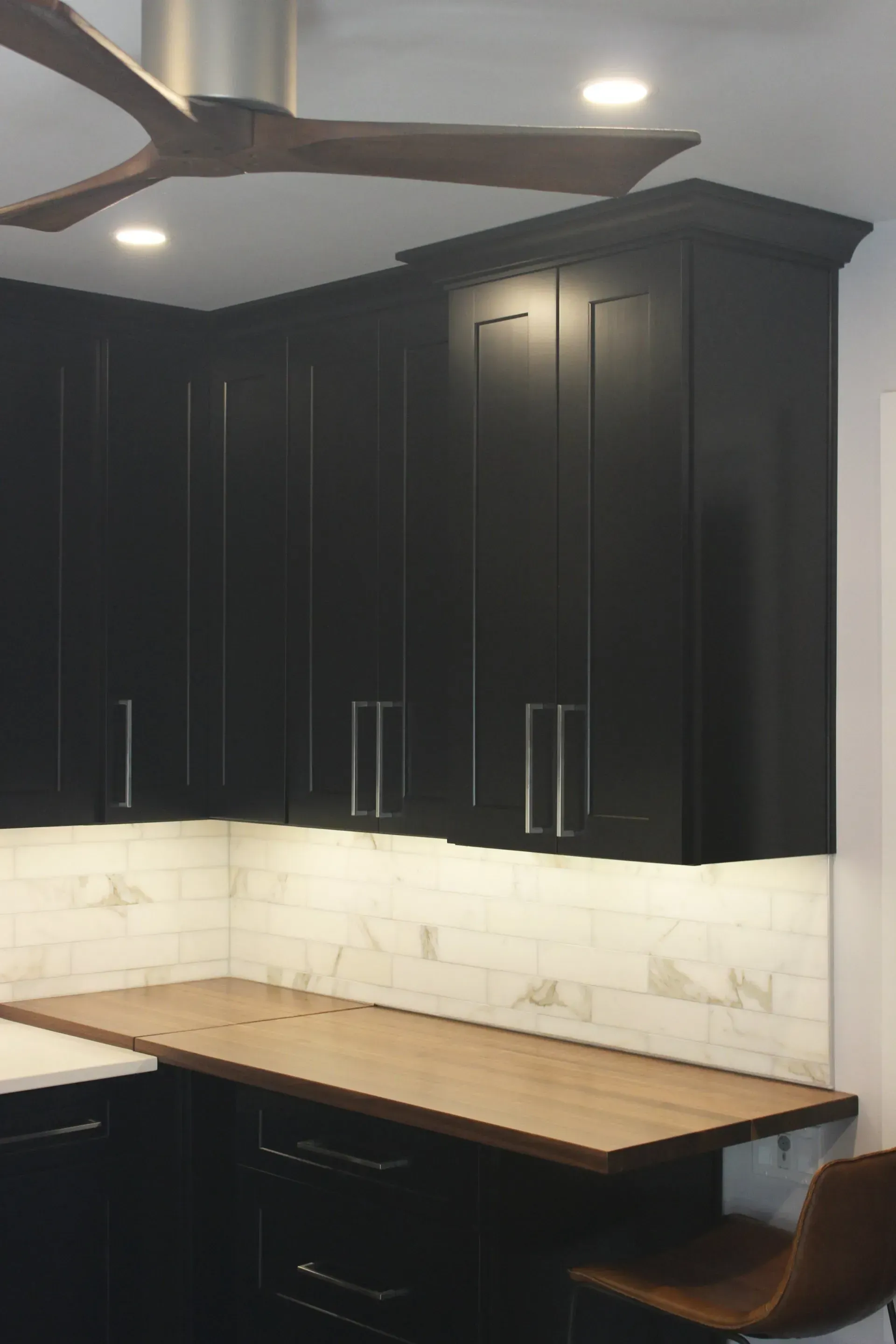 Black kitchen cabinets above a wood countertop with white tile backsplash and a brown chair in Bucks County PA