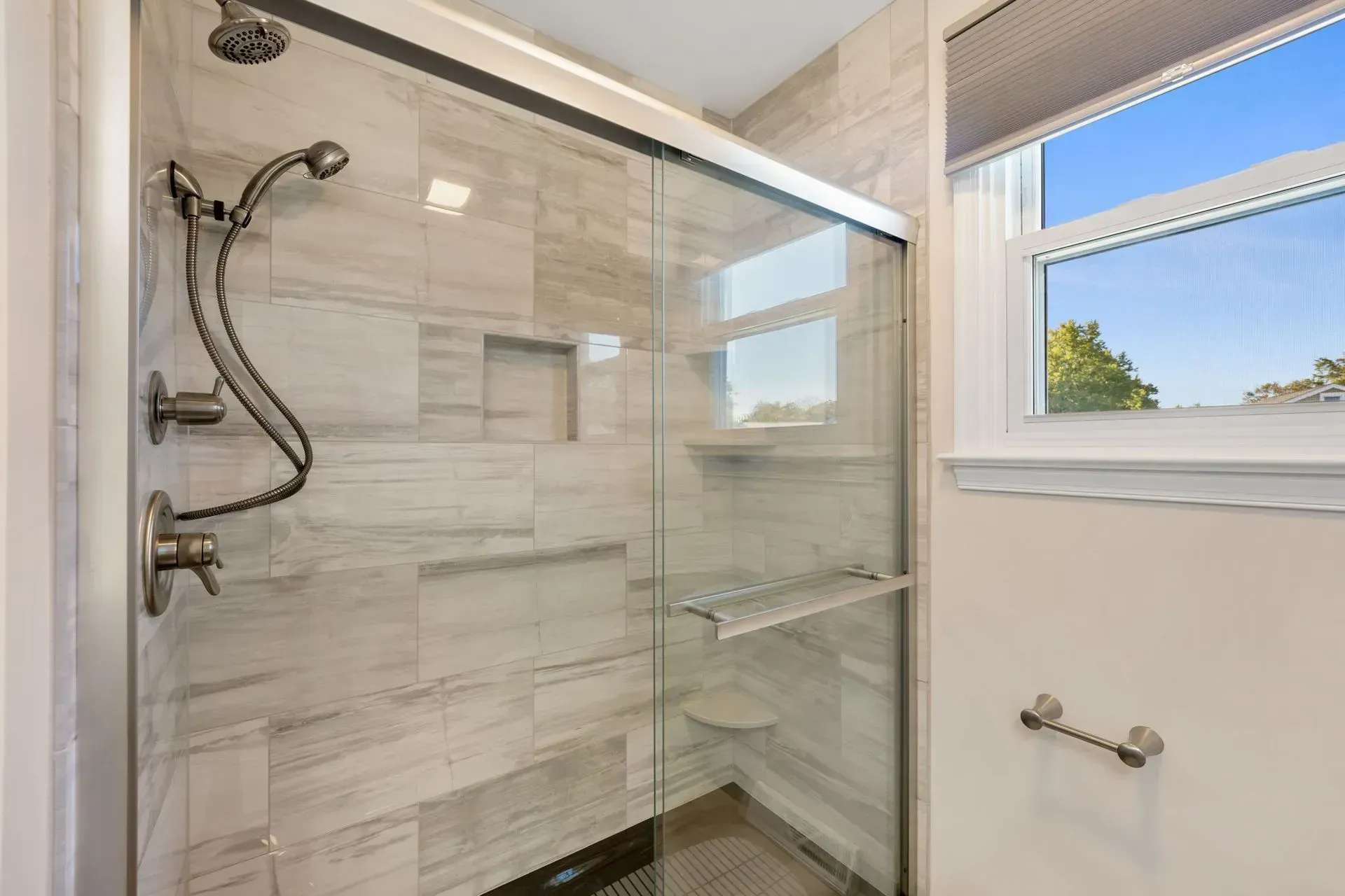 Modern tiled shower with glass door, chrome fixtures, and a window beside it In Bucks County PA