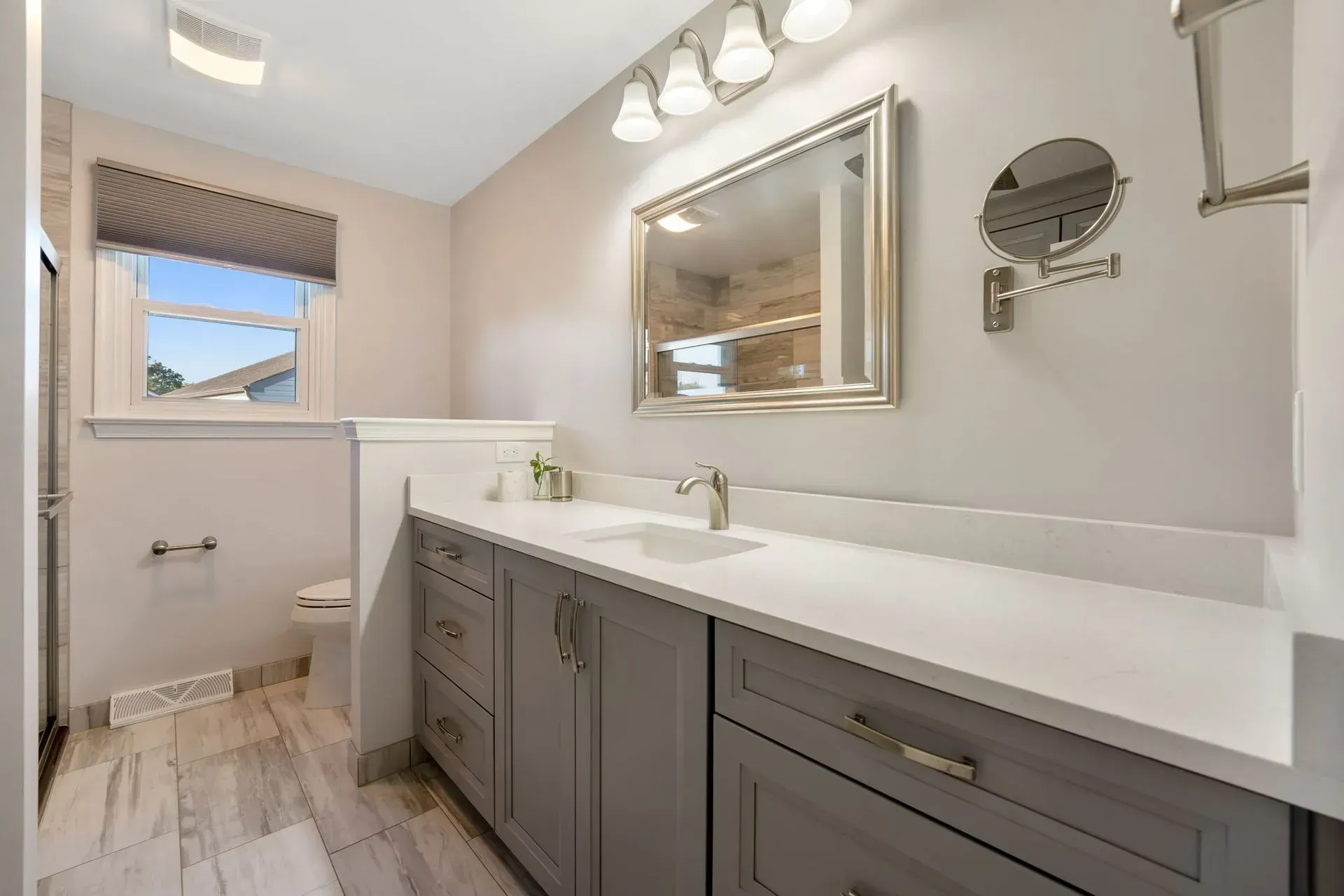 Bright bathroom with gray vanity, white countertop, large mirror, and window over a shower area In Bucks County PA