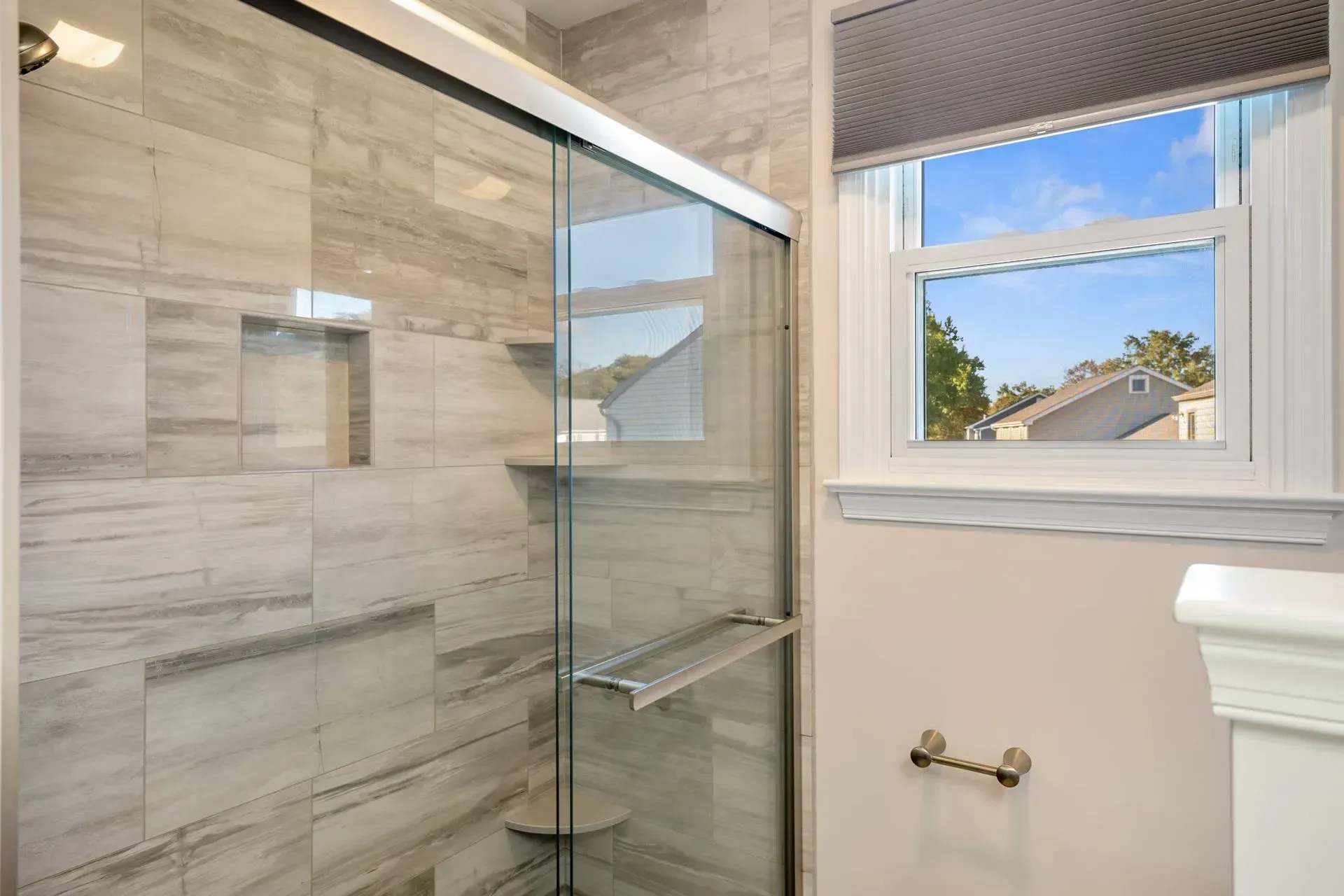 Modern bathroom with glass shower enclosure, beige tile walls, and a window above a towel bar In Bucks County PA