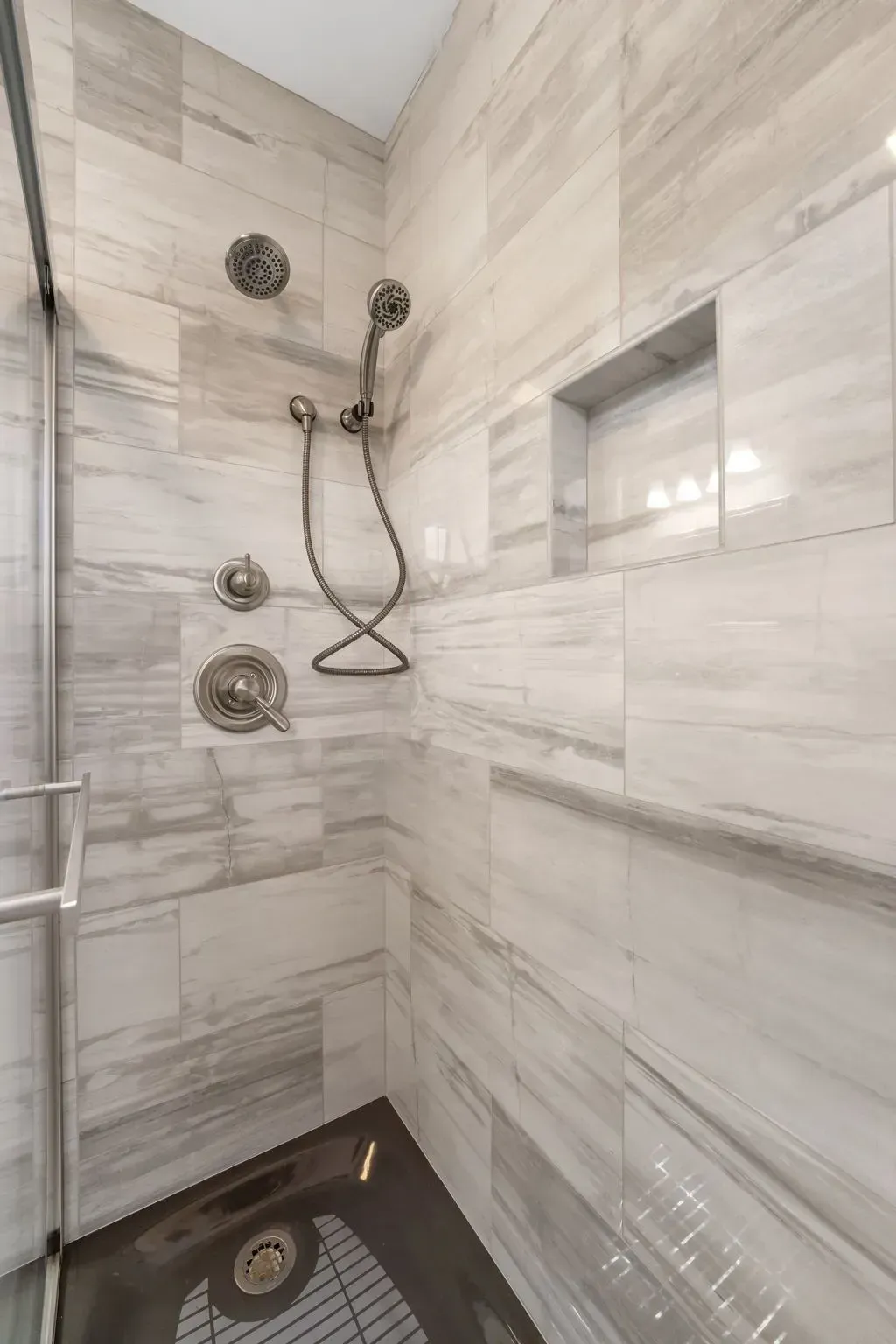 Modern tiled shower with chrome fixtures, corner shelf, and dark shower base In Bucks County PA
